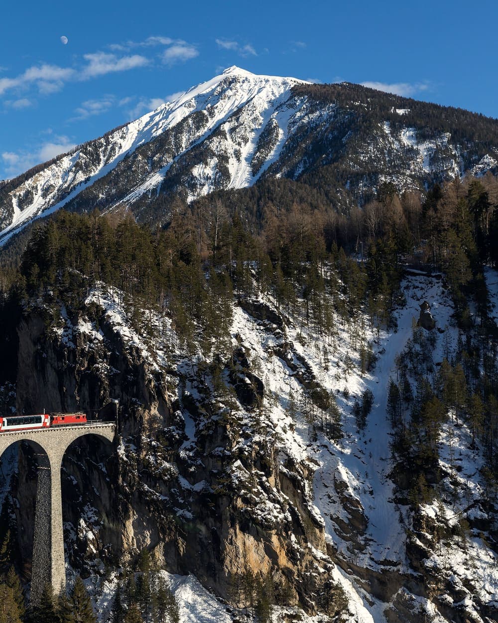 A red Swiss train crossing a viaduct through a snowy forest